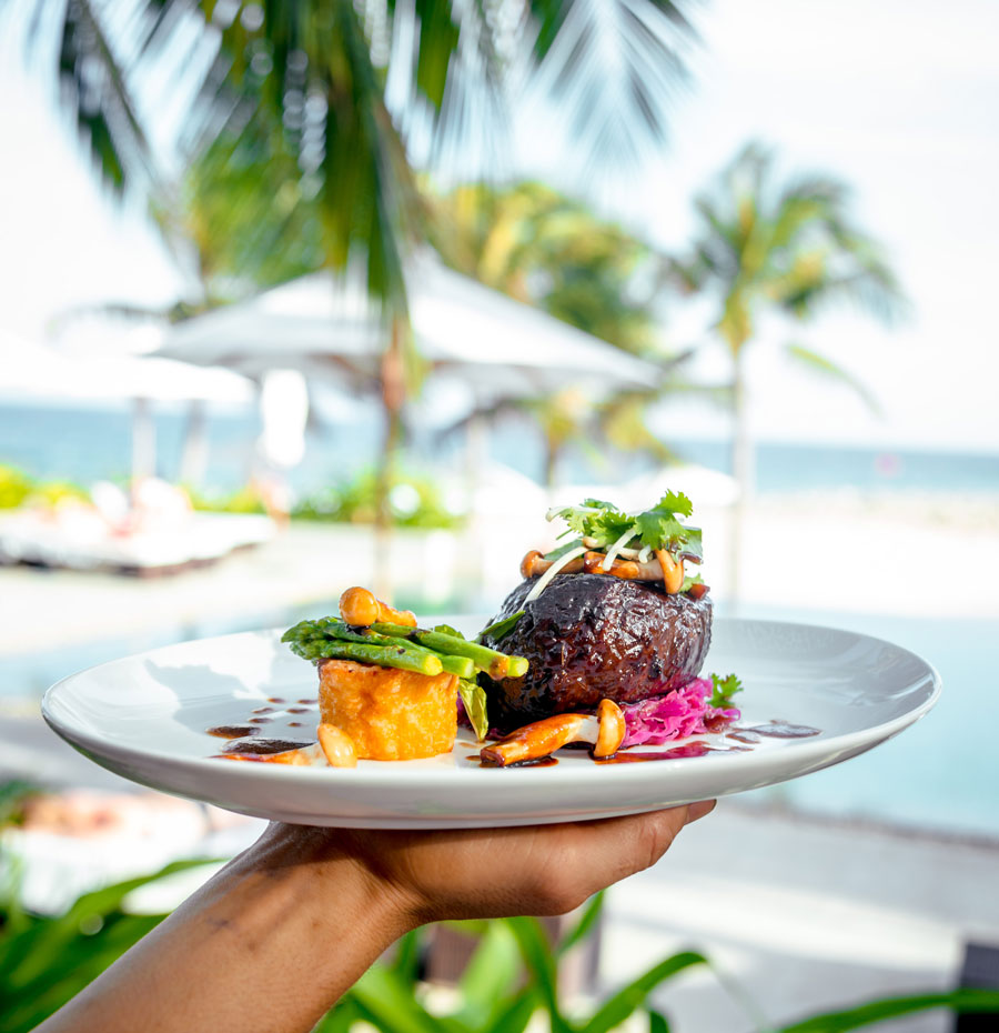 holding white plate with meat and potatoes near a beach